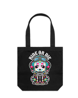 A black canvas tote bag with two handles, featuring a colorful graphic of a seated skeleton wearing motorbike helmet and goggles and the words 'RIDE OR DIE' printed above it, shown against a plain white background.