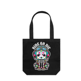 A black canvas tote bag with two handles, featuring a colorful graphic of a seated skeleton wearing motorbike helmet and goggles and the words 'RIDE OR DIE' printed above it, shown against a plain white background.