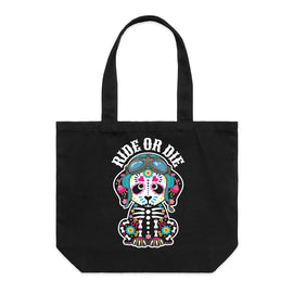 A black canvas tote bag with two handles, featuring a colorful graphic of a seated skeleton wearing motorbike helmet and goggles and the words 'RIDE OR DIE' printed above it, shown against a plain white background.